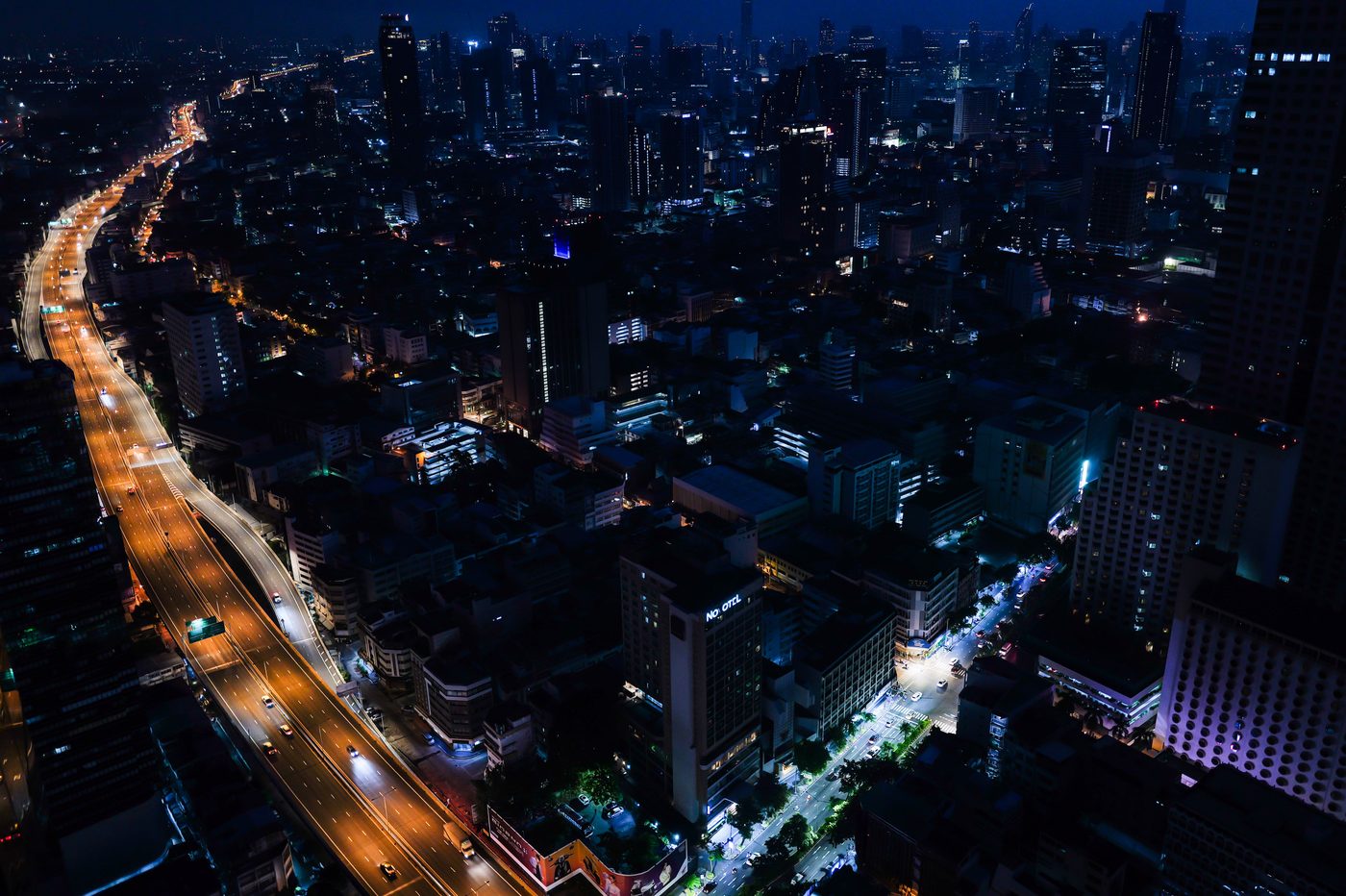 High-angle nighttime cityscape of illuminated buildings and a curving elevated highway filled with streetlights in Bangkok.