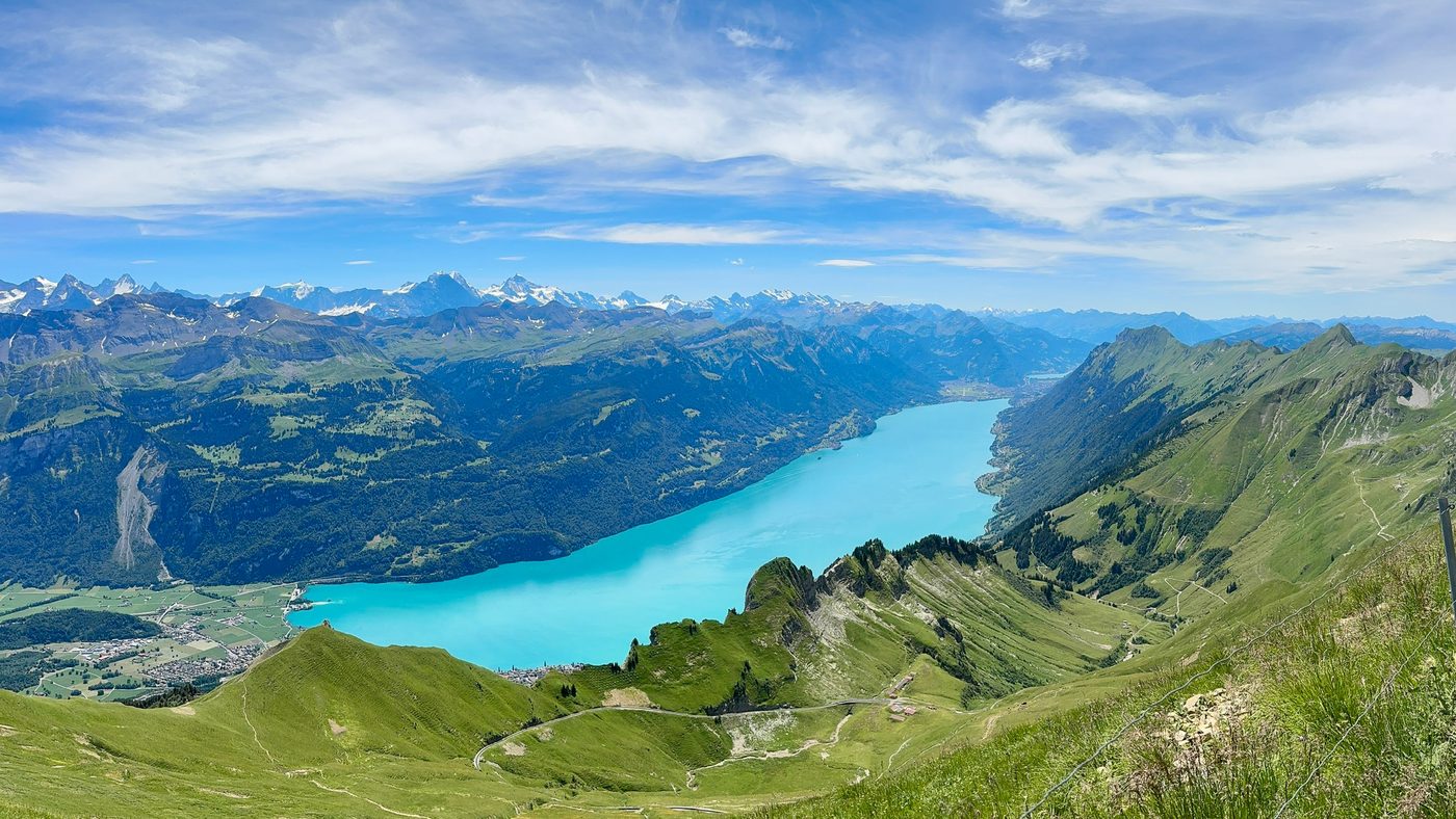 Panoramic mountain view over turquoise Lake Brienz in a valley surrounded by green slopes, with snow-capped alpine peaks in the distance.