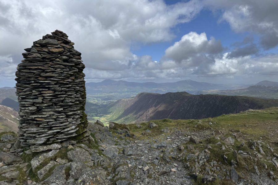Tall beehive-shaped stone cairn on a rocky mountain summit overlooking a deep green valley and distant mountains under a blue sky.