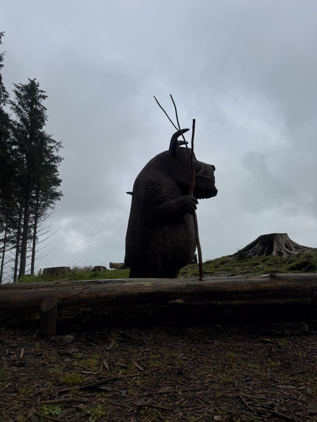 A wooden carved statue of the Gruffalo holding a tall stick stands behind a fallen log in a forest clearing.