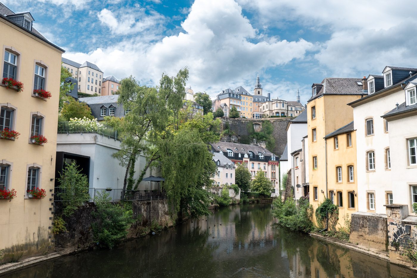 A river flanked by traditional European stone houses and green trees, with church spires and buildings atop high cliff walls in the background under a blue sky.
