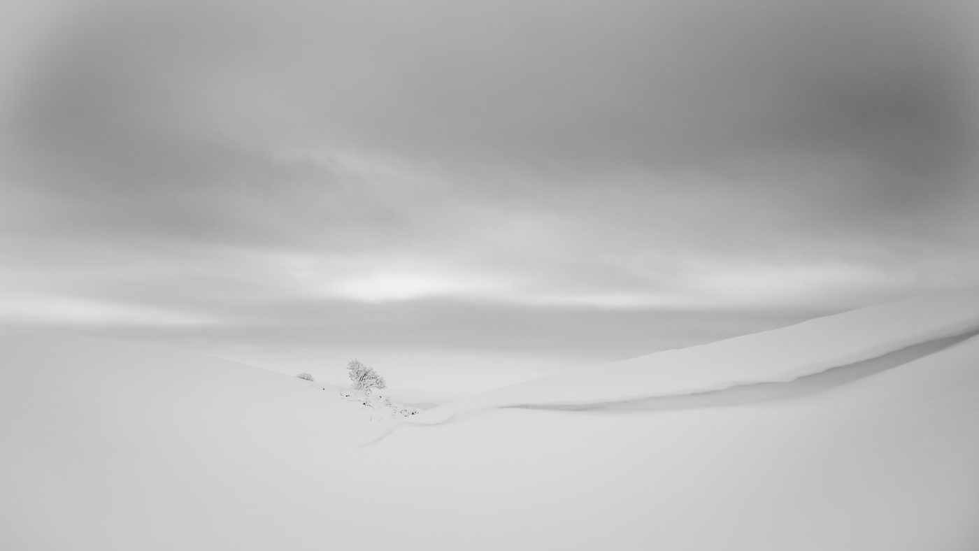 Deep snowdrifts form a sharp ridge curve under an overcast grey sky on a mountain slope.