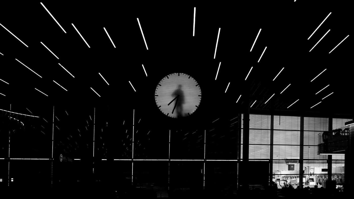 High-contrast view of a large circular backlit clock showing a human silhouette moving the clock hands, beneath radial linear ceiling lights.