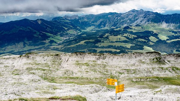 Yellow Swiss hiking signposts on rocky, light-grey karst terrain looking down over green valleys and mountain ridges under a cloudy sky.