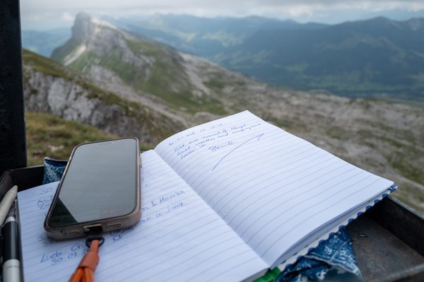 An open lined notebook with handwritten entries and a smartphone on it, with rugged limestone mountain ridges visible in the background.