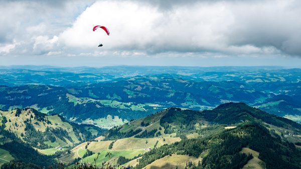 A paraglider with a red and white canopy glides over vast green rolling hills and valleys beneath large white clouds.