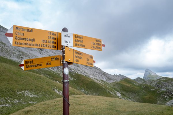 A yellow metal hiking trail signpost labeled Matten 1962 m with directions to various alpine destinations against a grassy and rocky mountain slope.