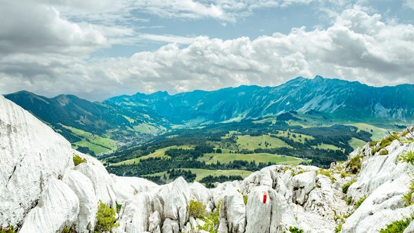 Heavily eroded white karst rocks with a red-and-white trail marker in the foreground overlooking green valleys and mountain ridges.