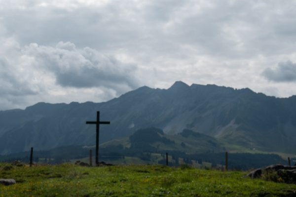 A wooden cross stands on a green grassy hill in the foreground with a mountain ridge and cloudy sky in the background.