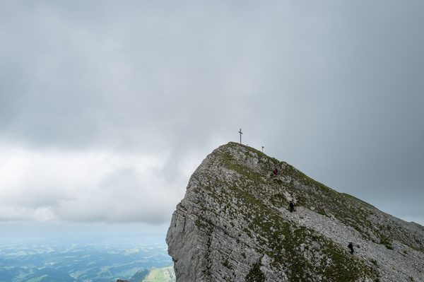 A rocky mountain ridge with a summit cross at the top, where several hikers are climbing under a cloudy sky.